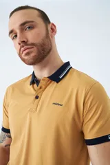 Remera tipo polo color ocre, de corte clásico, con cuello y mangas con terminaciones a contratono en azul marino y blanco.