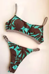 Bikini de dos piezas con estampado de hojas tropicales en marrón oscuro sobre fondo celeste turquesa. El corpiño es tipo top con tirantes finos y elástico a rayas blancas y negras en el contorno inferior. La bombacha es tipo colaless con elástico a rayas blancas y negras en los laterales.
