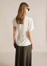 Blusa blanca de tejido fluido con textura, manga corta, cuello alto envolvente y cierre de cremallera en la parte posterior.