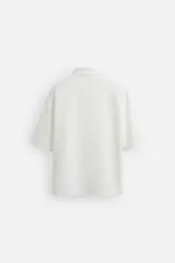 Sobrecamisa color beige de tejido elástico, con cuello solapa, manga corta y bolsillos de parche en el pecho. Cierre frontal con botones a presión.
