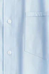 Camisa celeste de algodón suave, de corte holgado, con cuello clásico, botones frontales, canesú en la espalda, hombros caídos, bolsillo de parche en el pecho, mangas largas con puños abotonados y bajo redondeado.