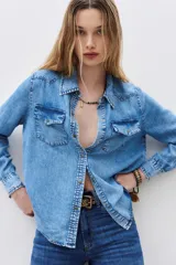 Camisa de denim celeste con cuello clásico, cierre frontal con botones a presión, dos bolsillos con solapa en el pecho y mangas largas con puños abotonados.