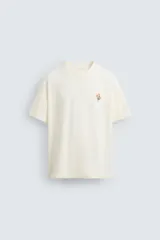 Camiseta color crudo de corte relaxed fit, con cuello redondo y mangas cortas. Presenta un pequeño bordado de flores en el pecho y un estampado fotográfico en la espalda.