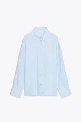Camisa celeste de lino, corte amplio, con cuello solapa, manga larga, bolsillo de plastrón en el delantero, bajo asimétrico y cierre frontal con botones.