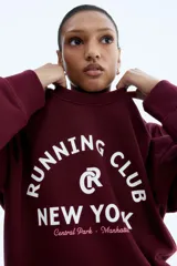 Buzo de tejido de punto color bordó, con cuello redondo y puños y bajo acanalados. Presenta un estampado frontal en blanco con el texto "RUNNING CLUB NEW YORK Central Park - Manhattan".