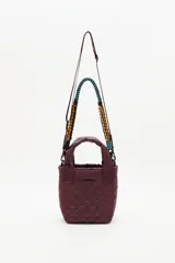 Cartera tipo tote mini acolchada color vino tinto (bordó oscuro), con textura matelassé. Posee dos asas cortas y una correa larga ajustable y desmontable con detalle de tejido trenzado multicolor (azul, negro, amarillo y bordó).
