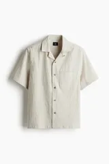 Camisa de manga corta color beige claro, confeccionada en tejido liviano de lino y algodón. Tiene cuello resort, tapeta lisa, bolsillo superior y bajo recto con aberturas laterales.