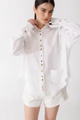 Camisa blanca de lino con botones de madera con forma de frutas, manga larga y corte holgado. Tiene un bolsillo en el pecho y bordado rojo con la frase "la vedetina".