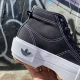 Zapatillas Adidas Nizza Trek de caña alta, color negro con costuras blancas contrastantes. Presentan suela de goma blanca con detalles en marrón y logo de Adidas en la lengüeta y el lateral.
