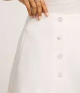 Short pollera de sastrería color blanco, con cintura alta y diseño de traspase frontal abotonado con botones forrados.