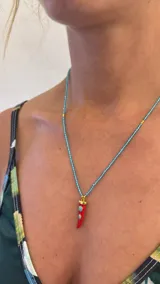 Collar de mostacillas de acrílico color turquesa con un dije colgante en forma de cuerno italiano (cornicello) rojo con incrustaciones de piedras turquesas. El engarce del dije y las cuentas separadoras son de metal dorado.