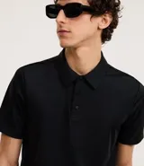 Camisa polo negra de manga corta, confeccionada en poliamida y elastano, con cuello inglés abotonado y tecnología Performance que proporciona mayor comodidad térmica, elasticidad y secado rápido.