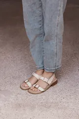 Sandalia plana de cuero color beige claro, con dos tiras anchas ajustables con hebillas metálicas cuadradas de color dorado. El borde de la plantilla está decorado con tachas metálicas doradas.