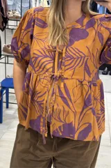 Blusa naranja con estampado de hojas color violeta, de calce holgado, escote en V con doble lazo ajustable y mangas 3/4 con volumen.