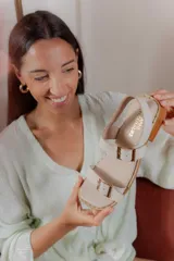 Sandalia plana de cuero color beige claro, con dos tiras anchas ajustables con hebillas metálicas cuadradas de color dorado. El borde de la plantilla está decorado con tachas metálicas doradas.