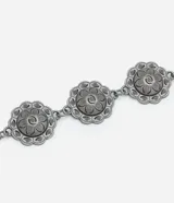 Cinturón de metal con acabado oscuro, compuesto por una cadena gruesa y una serie de dijes circulares con diseño floral en relieve. El cierre es de mosquetón con un dije floral como terminación.