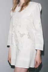 Vestido corto de algodón y lino color blanco, con cuello babero cuadrado, manga larga y bordados florales. Cierre en la espalda con abertura y botón.
