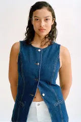 Chaleco largo de denim azul, con cuello redondo, cierre frontal con botones metálicos hasta la cintura y abertura inferior. Presenta bolsillos delanteros insertados y costuras decorativas.