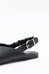Zapato destalonado de punta fina, color negro, con base baja y tira trasera elástica con hebilla metálica ajustable. Diseño minimalista.