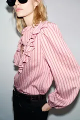 Camisa de manga larga color rosa claro con finas rayas verticales en tono rosado más oscuro. Presenta cuello mao y un gran volante fruncido que recorre el frente, desde los hombros hasta el centro. Los puños son elásticos.
