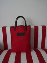 Bolso tipo tote bag de lona color rojo con detalles en cuero marrón en asas, borde superior y etiqueta frontal con logo.