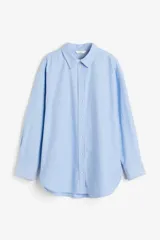 Camisa celeste de algodón Oxford, corte relajado, con cuello clásico, botones frontales, canesú con trabilla en la espalda, hombros caídos, mangas largas con puños sencillos, bolsillos superiores con solapa y bajo redondeado con espalda más larga.