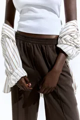 Pantalón marrón de corte holgado en sarga de mezcla de lyocell. Modelo de tiro alto con cintura fruncida, cierre decorativo y bolsillos laterales discretos. Piernas rectas con pliegues cosidos adelante y raya planchada atrás.