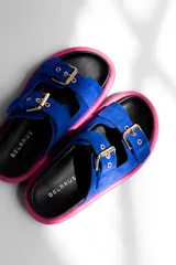 Sandalias planas de cuero gamuzado color azul eléctrico, con plantilla negra con el logo de la marca en dorado, borde fucsia y hebillas doradas ajustables.