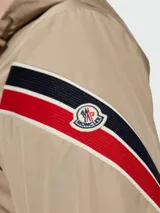 Rompevientos beige con capucha y cierre frontal con cremallera. Tiene bolsillos laterales con cierre de botón a presión y detalle de franjas rojas, blancas y negras en la manga izquierda.