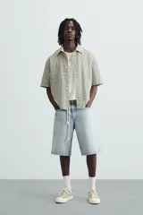 Bermuda de denim de algodón, color gris claro lavado, de corte ancho (baggy fit) y largo hasta la rodilla. Presenta cinco bolsillos y cierre frontal con cremallera y botón.