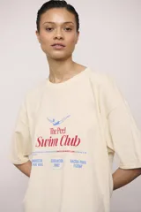 Remerón de manga corta color crema, corte oversize y cuello redondo. Estampado frontal con texto y un dibujo de una persona nadando, con la frase "The Peel Swim Club".