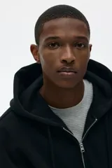 Campera con capucha y cierre frontal, color negro, confeccionada en tejido pesado de mezcla de algodón con interior cepillado suave. Presenta hombros caídos, capucha forrada con cordón ajustable y bolsillos delanteros tipo canguro. Puños y bajo acanalados.