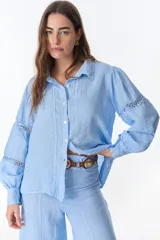 Camisa celeste de lino con cuello clásico, abotonada al frente y mangas largas con detalle de puntilla en los hombros.