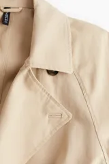Gabardina larga color beige de corte holgado, con solapas de muesca, mangas raglán, bolsillos laterales y cinturón para atar.
