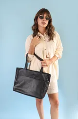 Cartera tipo tote de cuero graneado color negro, con textura uniforme y suave al tacto. Tiene manijas de 55 cm, bolsillo frontal con broche imantado, bolsillo trasero con cierre metálico, cierre principal metálico, forro interior y tres bolsillos internos (uno con cierre, uno plaqué y uno tipo sobre con solapa y botón).