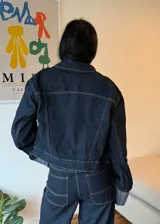 Campera de jean azul oscuro con costuras en contraste color crema, corte cropped, cuello clásico, cierre frontal con botones metálicos plateados, bolsillos de parche en el pecho y mangas largas con puños anchos con dobladillo en color gris. 