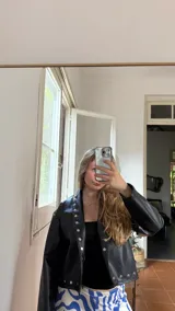 Campera corta de cuero sintético color marrón oscuro, con cuello solapa y detalles de tachas plateadas en el frente, hombros y puños. Presenta mangas amplias.