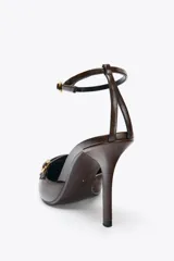 Zapato de tacón destalonado color marrón, con punta fina, tiras frontales con hebillas metálicas y pulsera al tobillo con cierre de hebilla.