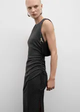 Vestido midi negro con estampado de lunares blancos, de tejido fluido y corte entallado. Presenta tirantes anchos, fruncido lateral con lazo, escote redondo y cierre de cremallera invisible en la parte posterior.