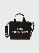 Bolso tote pequeño de lona color negro con doble asa de mano y correa de hombro ajustable y extraíble. Estampado frontal con el texto "THE TOTE BAG MARC JACOBS" en color blanco.
