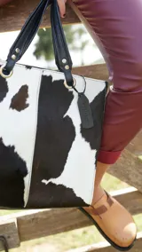 Bolso tipo tote de cuero nonato con estampado de vaca en blanco y negro, con asas de cuero negro y etiqueta colgante de cuero negro con el nombre de la marca.