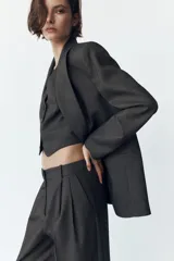 Chaleco corto gris oscuro con escote en pico, sin mangas, bolsillos de ojal y cierre frontal con botones.