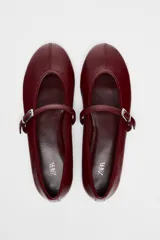 Zapato tipo bailarina color borgoña con efecto acharolado, acabado en punta redonda y tira con hebilla ajustable en la parte delantera.