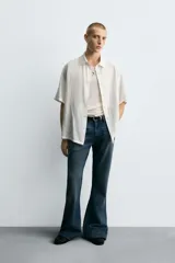 Camisa blanca de manga corta con cuello solapa y textura jacquard irregular. Corte relajado (relaxed fit) con aberturas laterales y cierre frontal de botonadura.