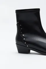 Bota corta de cuero sintético color marrón oscuro, con caña elástica y puntera afilada. Presenta una hilera de tachas metálicas doradas en el lateral exterior y un taco bajo y estable.