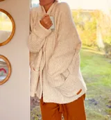 Cárdigan largo color crudo, de tejido de punto grueso con cuello alto, manga larga y bolsillos delanteros.