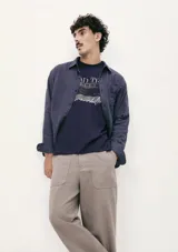 Camiseta blanca de algodón con cuello redondo y mangas cortas. Estampa rectangular con borde negro que contiene la frase "NATURAL ATMOSPHERE" en letras mayúsculas y una imagen de montañas y mar.