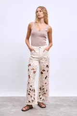 Pantalón de denim color crudo, corte ancho y tiro alto, con grandes calados florales bordados en contraste a lo largo de ambas piernas.