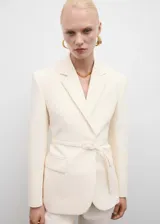 Blazer color crema con cuello de solapa, manga larga con puños abotonados, cinturón con hebilla y bolsillos delanteros con solapa.