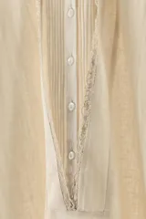 Kaftán color crema de hilatura con lino, cuello redondo y manga tres cuartos con bordados a contraste. Detalle de pliegues en delantero y acabados en puntilla a tono. Cierre frontal con botones.